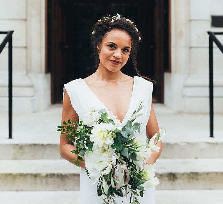 A bride stands in front of steps outside with halo braid and holding a bouquet