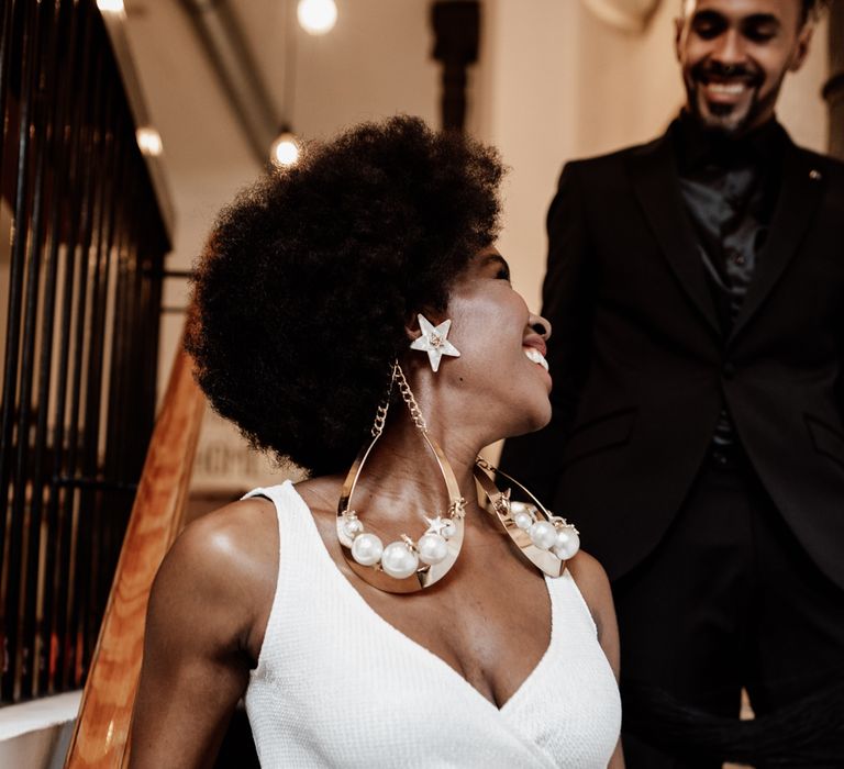 A Black bride wears over sized earrings. She looks back towards her groom as he walks down the stairs.