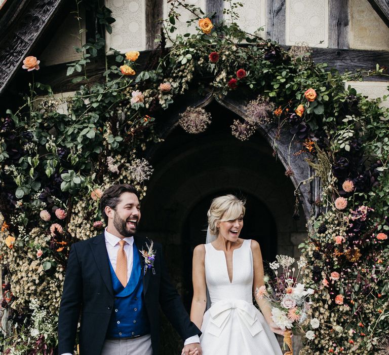 Blonde bride with a fringe in a Jesus Peiro wedding dress with V front and bow waist detail smiling as she exits the church with her new husband.