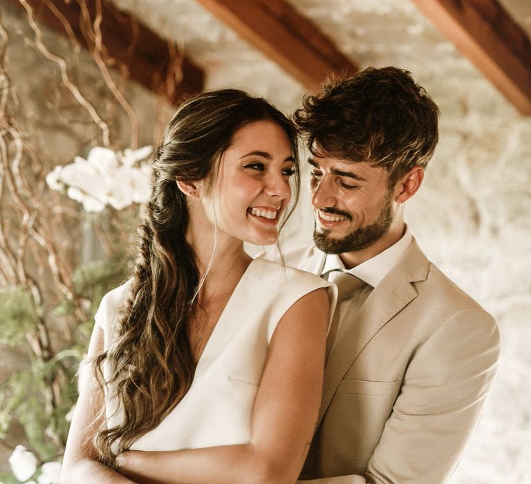 Bearded groom in a beige suit embracing his bride in separates with a long braided half up half down wedding hairstyle