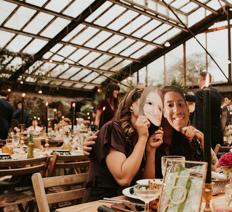 Wedding guests sitting at the autumn themed wedding tables holding up face masks of the groom 