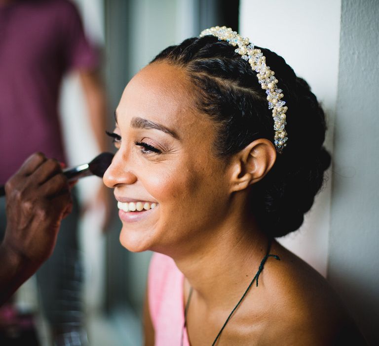 The bride smiling while having her blusher applied, she is wearing her hair in a braided up-do with a beaded headband