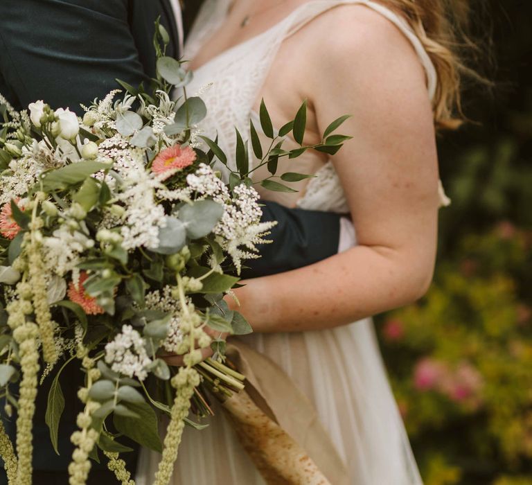 Bride & groom embrace whilst bride holds floral bouquet featuring whites and light pinks