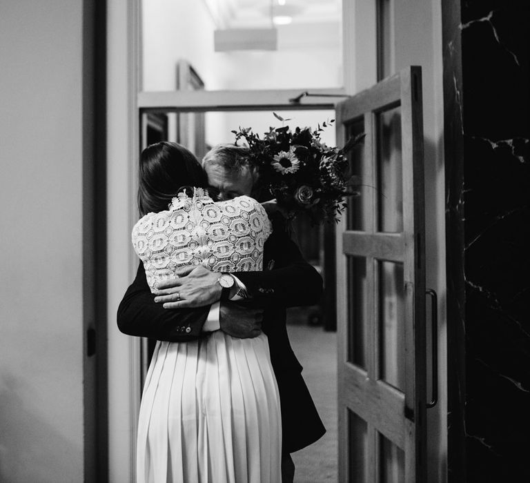 Bride in long sleeved lace top Self Portrait wedding dress holding colourful bouquet hugs groom in brown woollen blazer and waistcoat at Bristol Registry Office