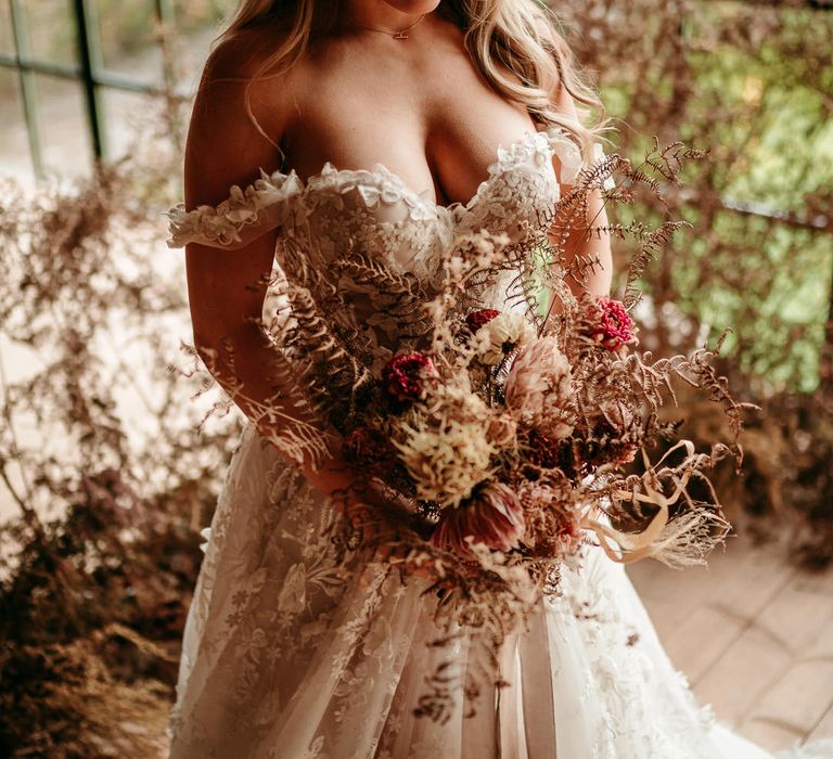 Bride in an off the shoulder wedding dress looking down at her dried flowers wedding bouquet with dahlias and grasses 