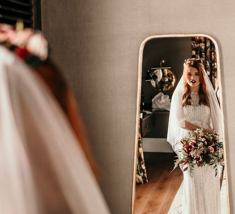 Bride looks in the mirror of the morning of her wedding whilst holding floral bouquet