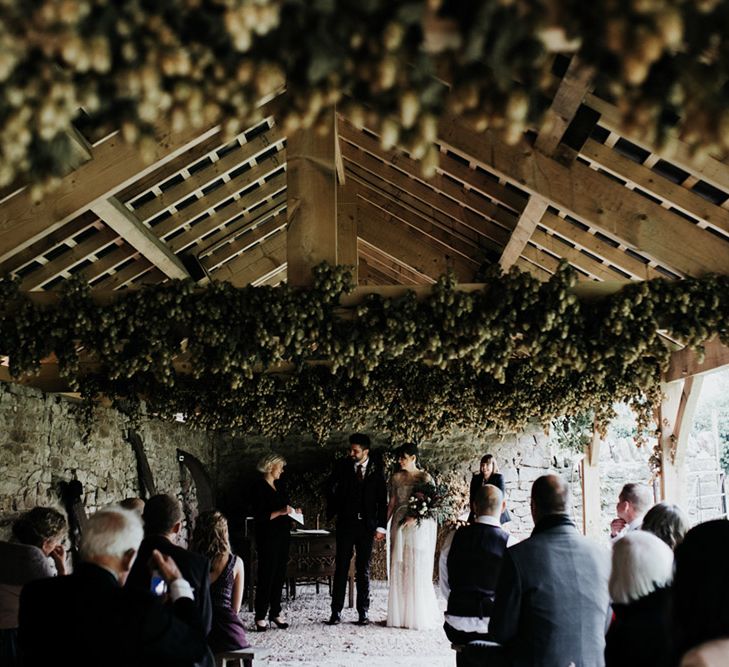 A bride and groom exit their civil ceremony. The venue is outside but under cover of beams and foliage. 