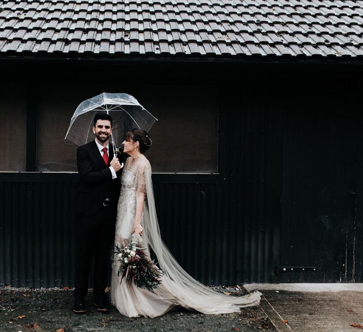 A bride and groom stand under an umbrella for their couples portraits. The bride holds a homemade wedding bouquet.