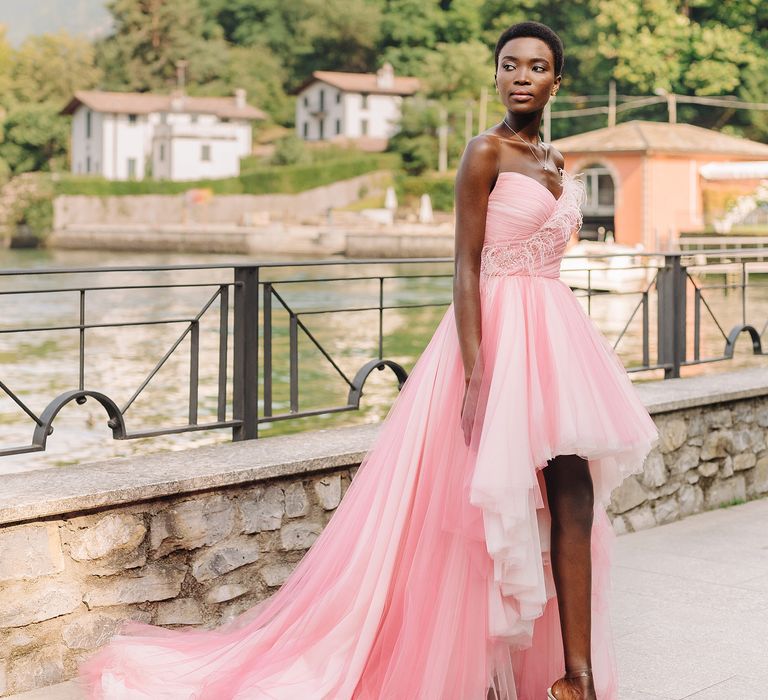 Black bride stands beside Lake Como whilst wearing pink bridal gown with feathering 