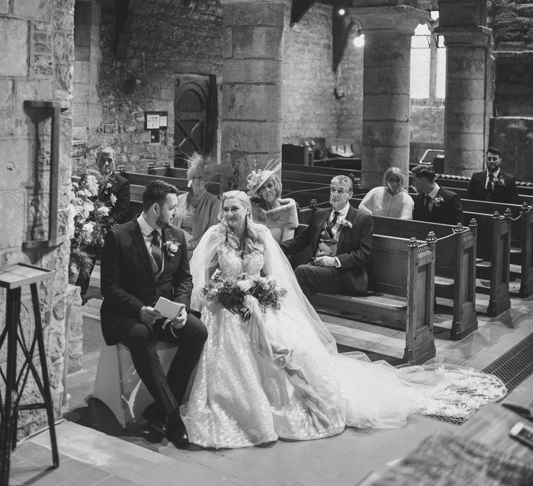 Bride in a princess wedding dress and bridal cape sitting at the altar with her groom in a navy suit 