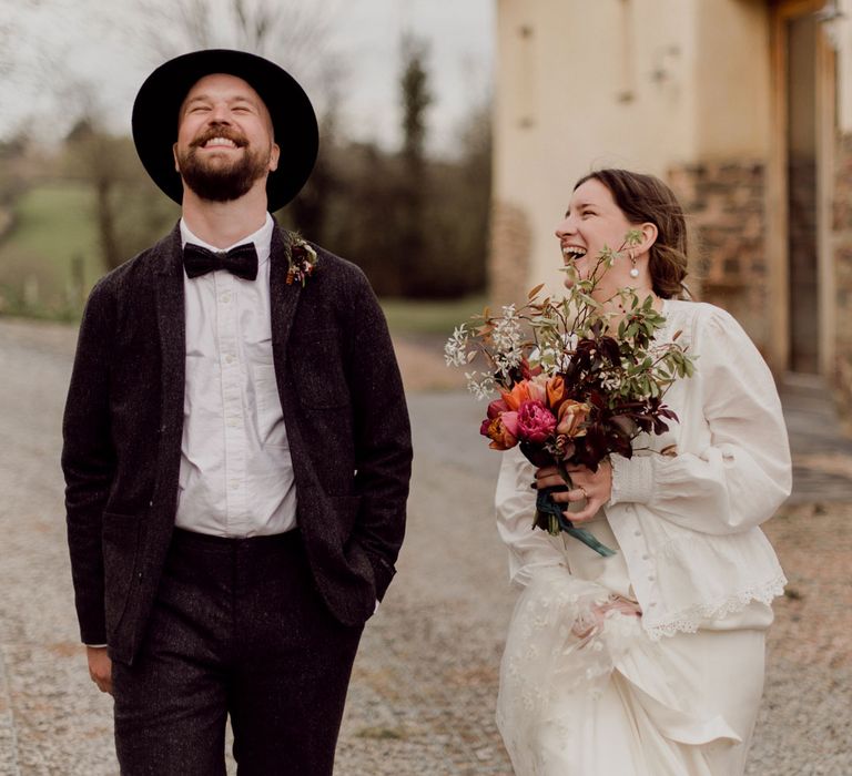 Laughing bride in white blouse, Charlie Brear wedding dress and applique veil holds multicoloured wedding bouquet whilst walking alongside laughing groom in grey tweed suit, bow tie and black fedora at garden party wedding in Devon