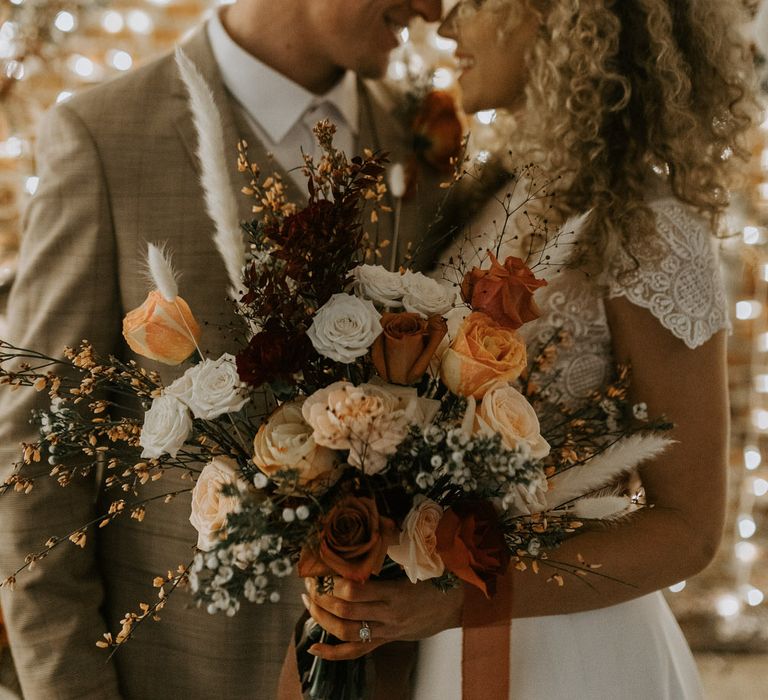 Bride and groom standing nose to nose in front of fairy lights, the bride is holding a large bouquet of mixed roses