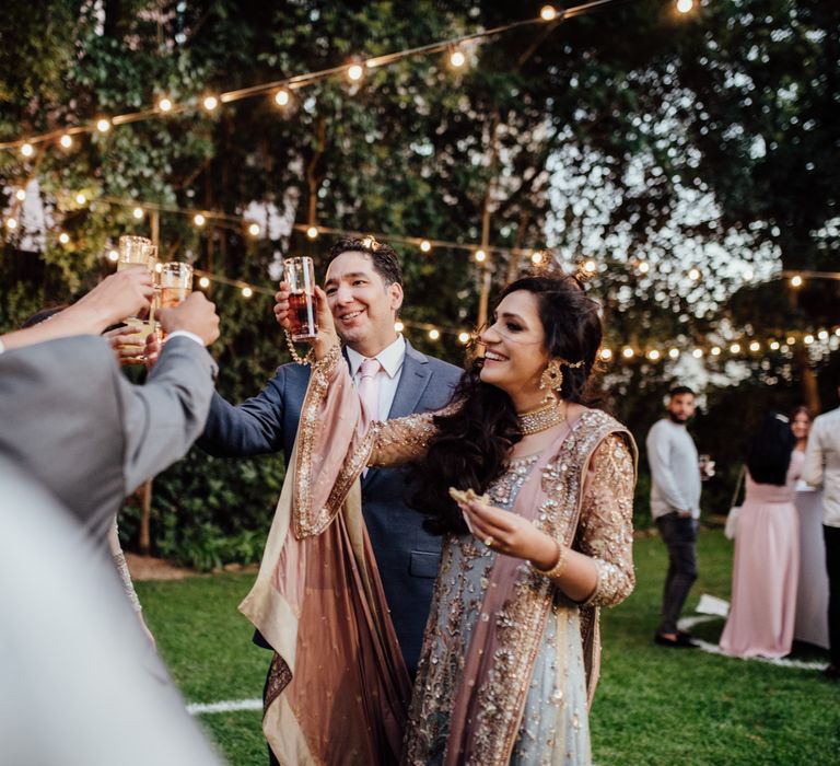 Bride lifts glass in celebration with groom as twinkling lights hang above