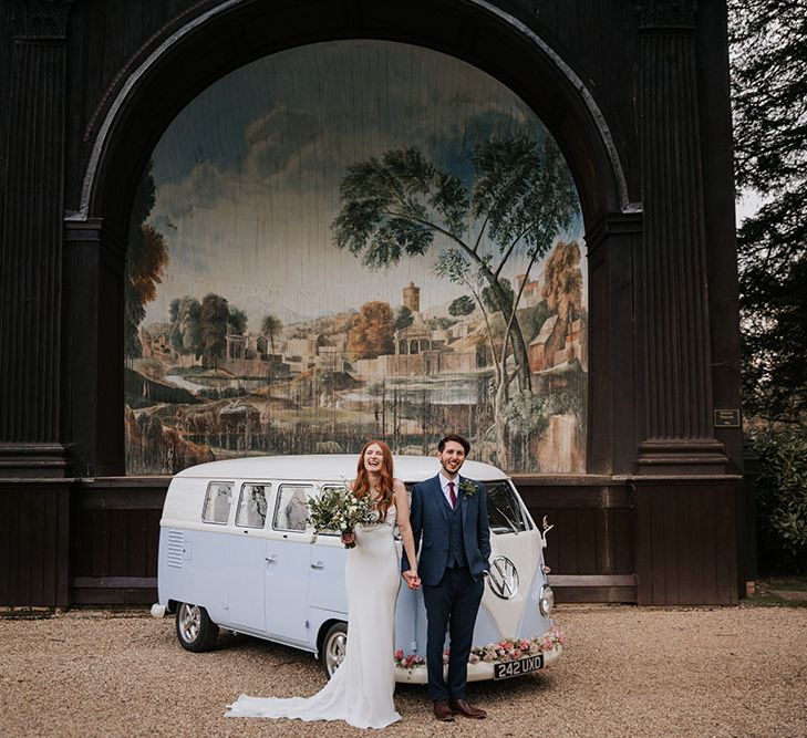 Bride and groom holding hands standing in front of their blue VW camper can and the outdoor singing theatre at Larmer Tree wedding venue