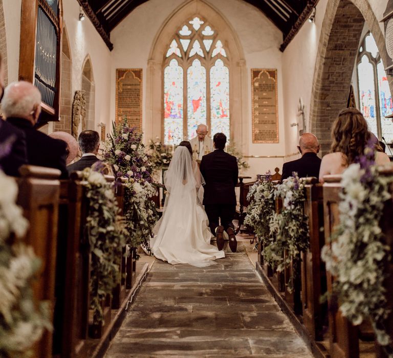 Interior of church wedding ceremony with bride in Elbeth Gillis gown and long veil kneels at the altar with groom in brown suit before vicar in white robes