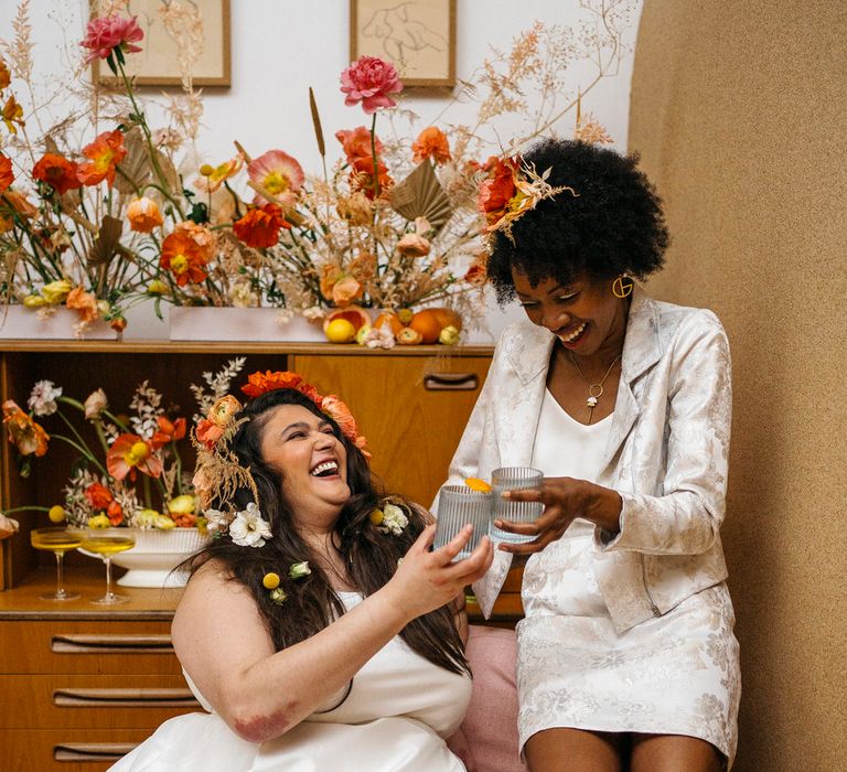 Two brides laughing in front of 70s wedding decor with floral display, both wearing bridal separates from Paper To Lace