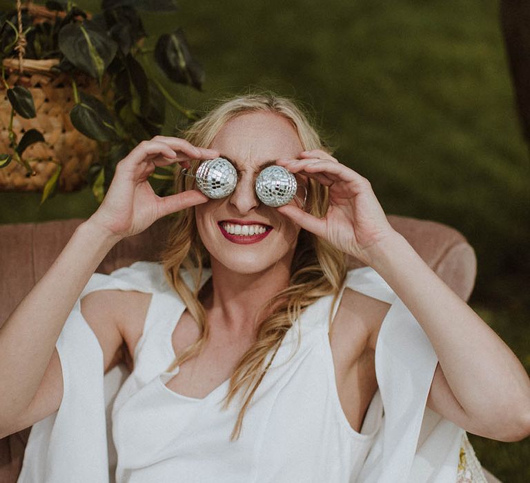 Bride in a jumpsuit and cape holding miniature disco balls at her eyes 