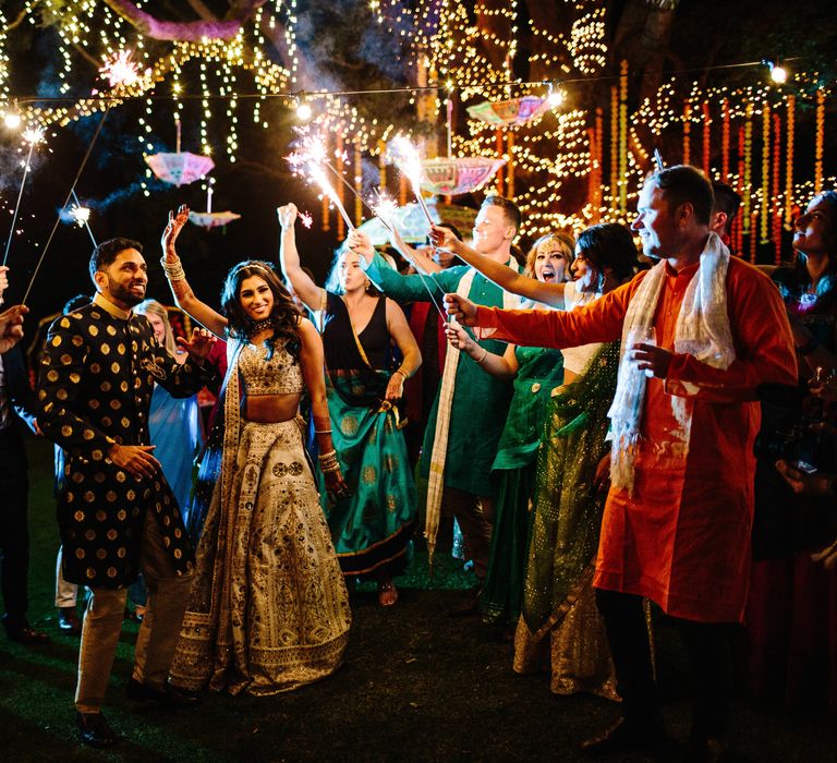 Bride & groom stand with wedding guests outdoors as they are surrounded by lights in the trees