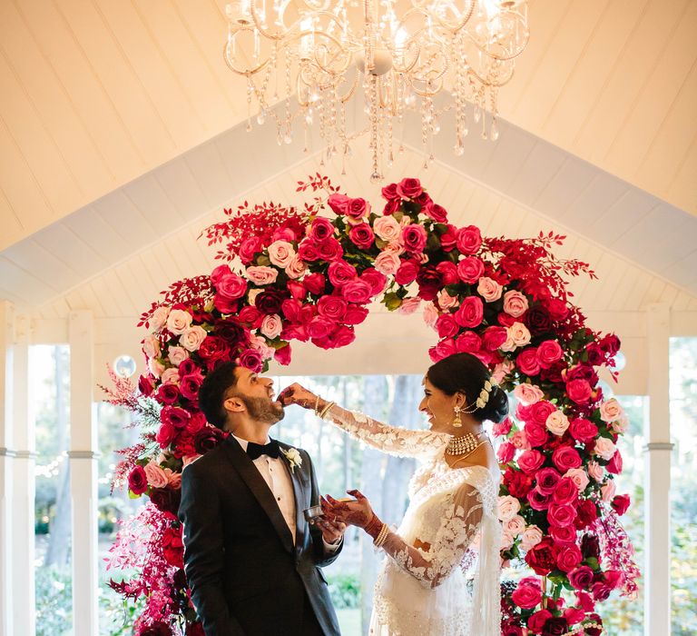 Groom holds brides hand as they stand below floral archway covered in bright red and pink flowers