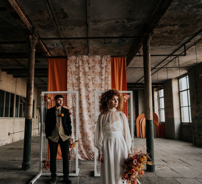 Bride and groom in a jumpsuit and patterned shirt in an industrial wedding venue with patterned fabric backdrop 