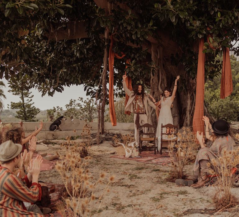 Bride & groom celebrate after marriage in hippie wedding under secular ficus tree complete with hanging orange fabric and outdoor wedding ceremony