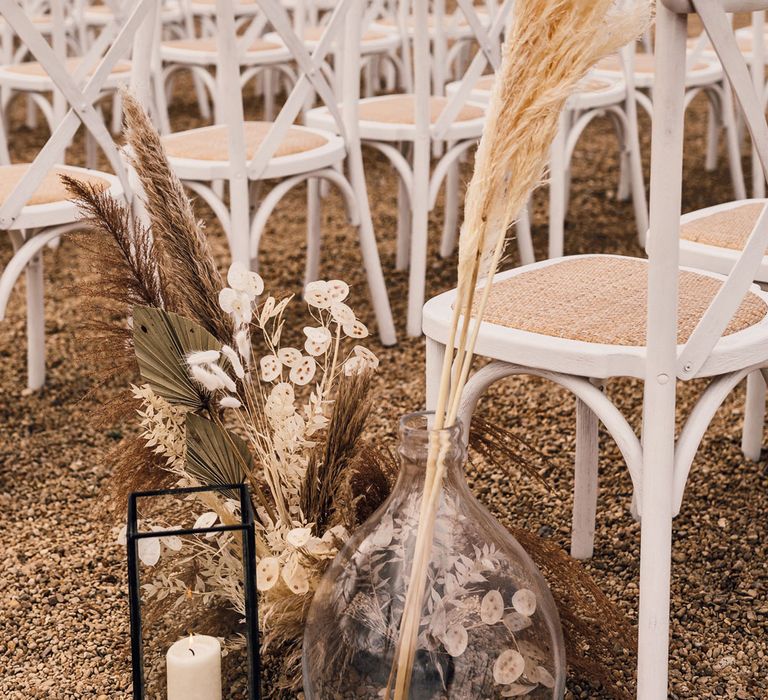 Candle in glass candleholder and dried flowers in glass vases in the aisle next to white wooden chairs for outdoor wedding ceremony in Dorset