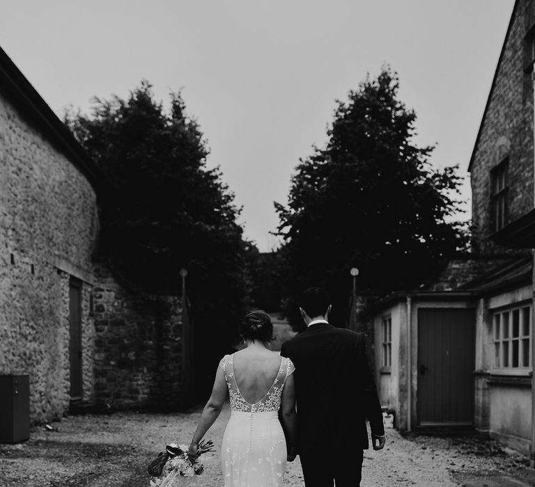 Bride in open back Rime Arodaky wedding dress holding dried bridal bouquet holds hands with groom in blue suit as they walk through courtyard at summer wedding in Dorset