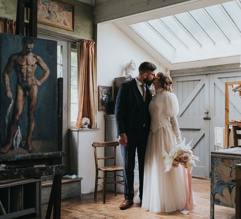 Bride & groom kiss on their wedding day as bride holds pastel bouquet with pampas grass and tied with pink ribbon