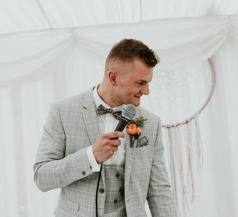 Groom in a check three-piece grey wedding suit and floral bow tie delivering his wedding speech during the marquee reception