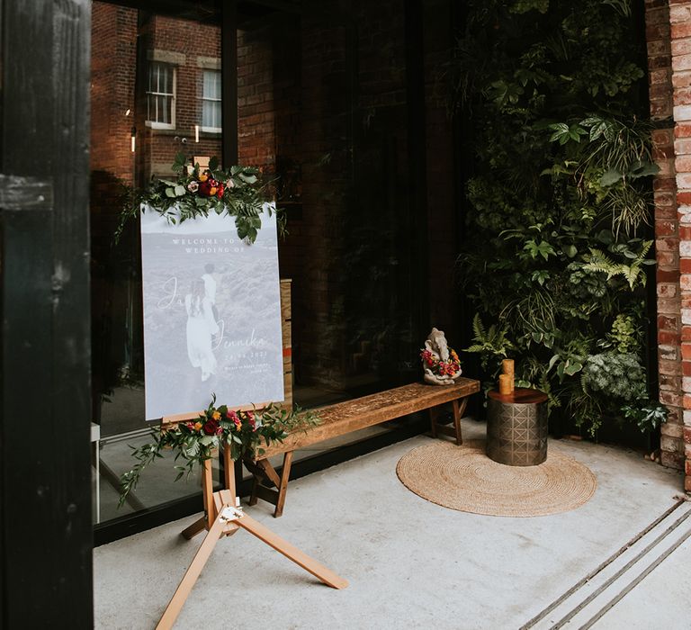 A welcome signs rests on an easel decorated with brightly coloured flowers to welcome guests to an Indian British wedding. 