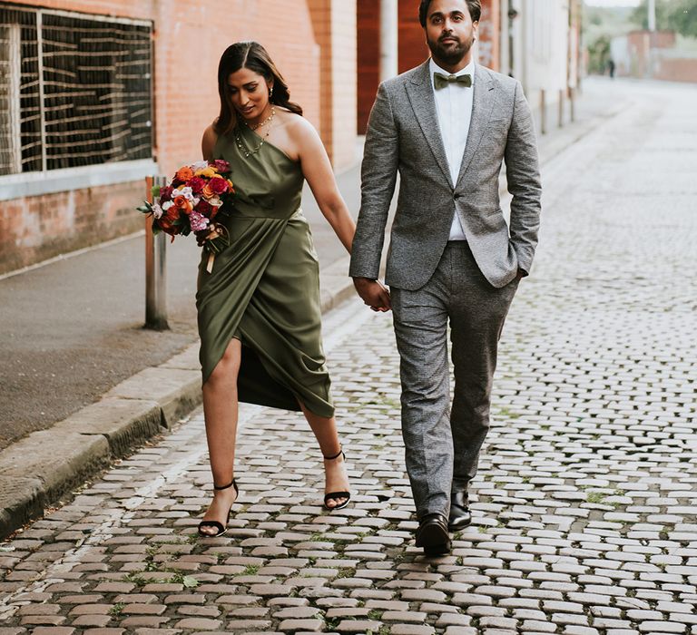 An Indian British couple walk to their wedding breakfast. He wears a grey suit and she wears and dark green dress.