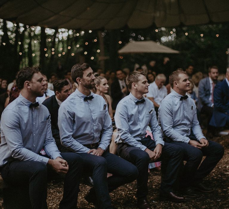 Groomsmen sit and watch ceremony outdoors as they wear pale blue shirts and bowties with no blazers