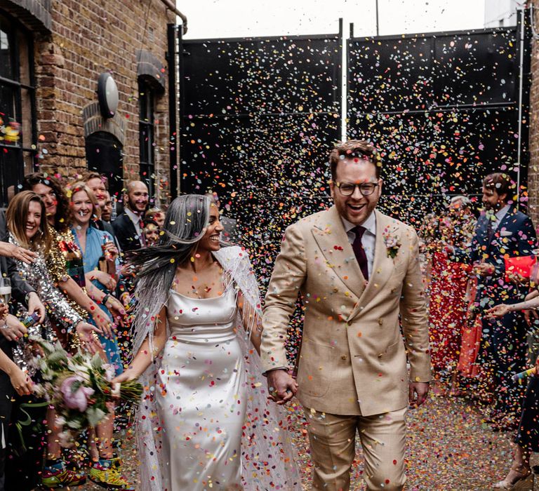 Bride in satin Halfpenny London wedding dress and tasselled bridal cape and groom in double breasted linen suit and gold Nike trainers walk through multicoloured confetti thrown by guests for wedding at Loft Studios London