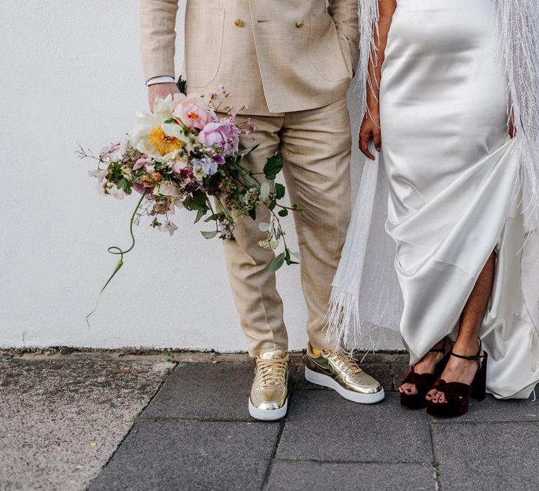 Bride in satin Halfpenny wedding dress and tasselled bridal cape stands holding up skirt to reveal velvet burgundy block heels next to groom in linen suit with gold Nike Air Force 1 trainers holding multicoloured wedding bouquet after wedding ceremony at Loft Studios London