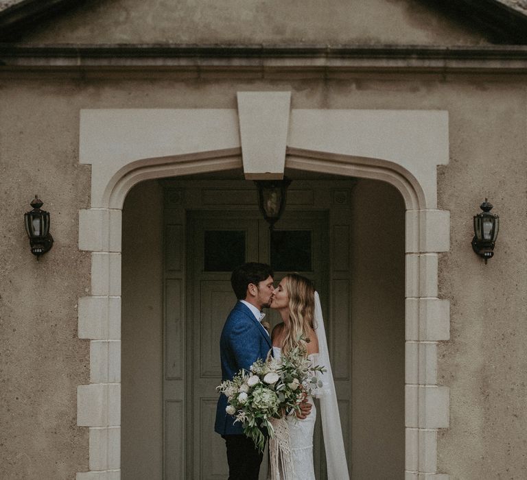 Bride in Grace Loves Lace bardot wedding dress and veil holding white and green bridal bouquet kisses groom in blue suit jacket in doorway at Isle of Wight wedding with macrame wedding decor