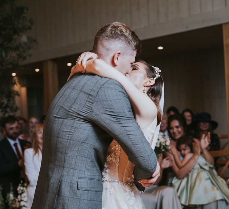 Bride in applique Berta wedding dress, headband and veil kisses groom in grey checked suit during wedding ceremony at Primrose Hill Farm