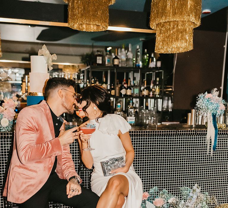 Stylish bride and groom sitting at the bar drinking cocktail in a pink velvet jacket and wedding dress with front split and boots 