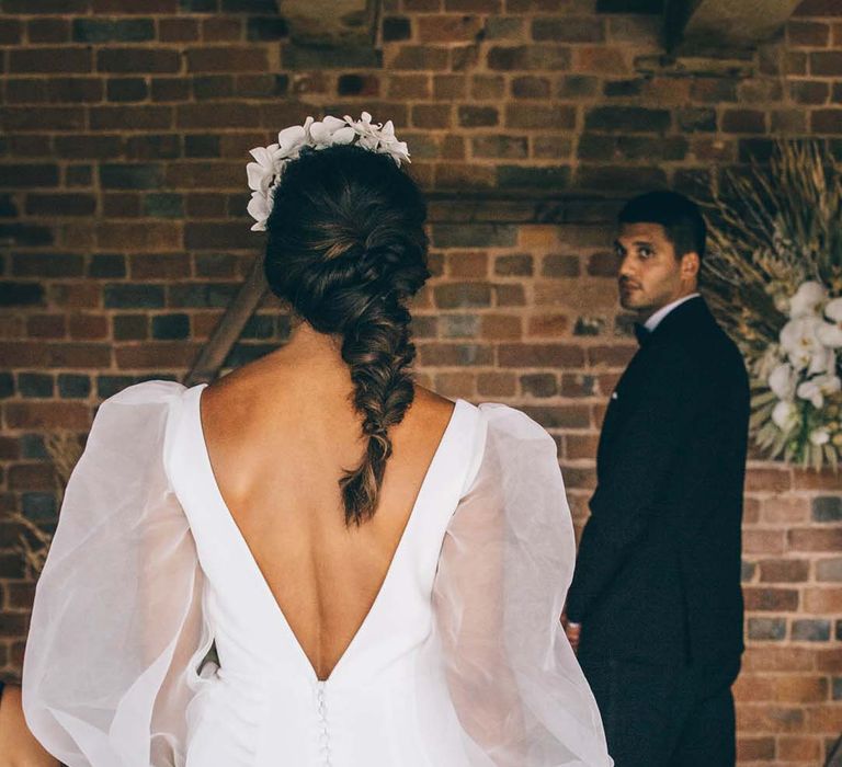 Bride in a fitted wedding dress with low V back, button detail and sheer long sleeves with a braided up do walking down the aisle 