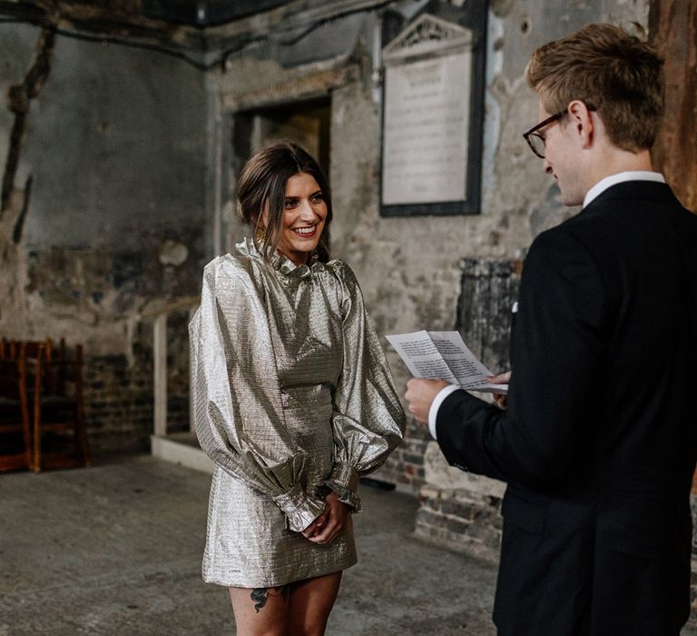 Tattooed bride stands in gold dress with Groom reading in dark suit in The Asylum London Chapel