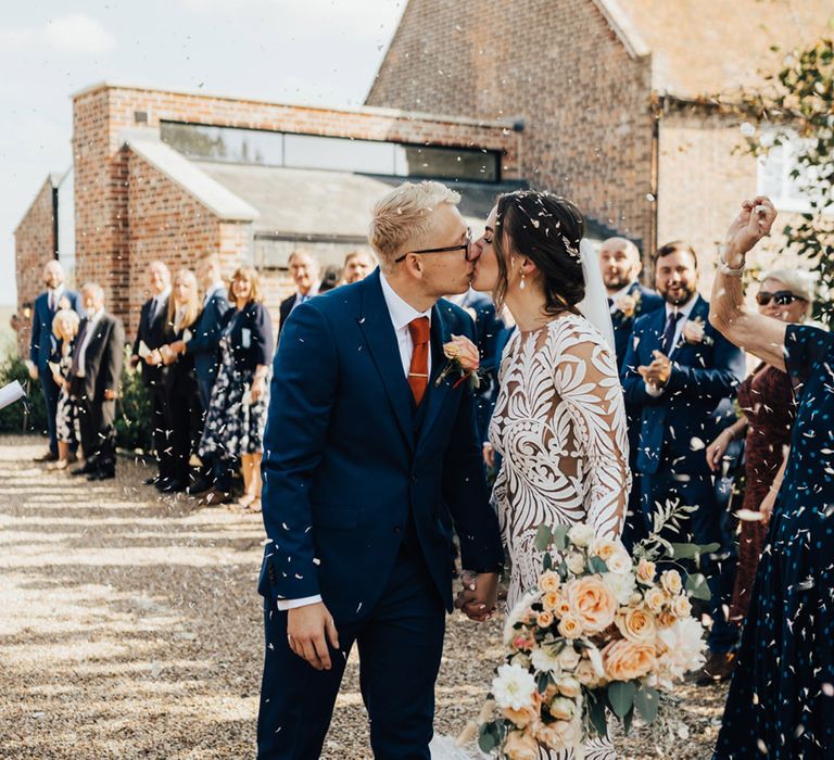 Bride with hair up do & Boho wedding dress kisses Groom in three piece navy suit with white shirt at Elmley Nature Reserve