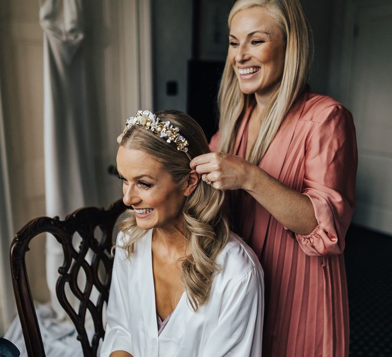 Bride in white satin dressing gown and curled hair has bespoke floral headband placed by woman in pink stain dress before church wedding ceremony