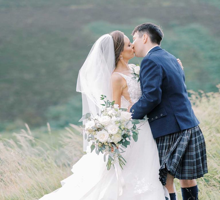 Groom in a tartan kilt and blue wool jacket kissing his bride in a princess wedding dress at their fairytale wedding 