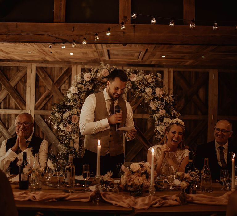 Groom in a waistcoat giving his wedding speech at Dewsall Court wedding with floral arch behind the top table 