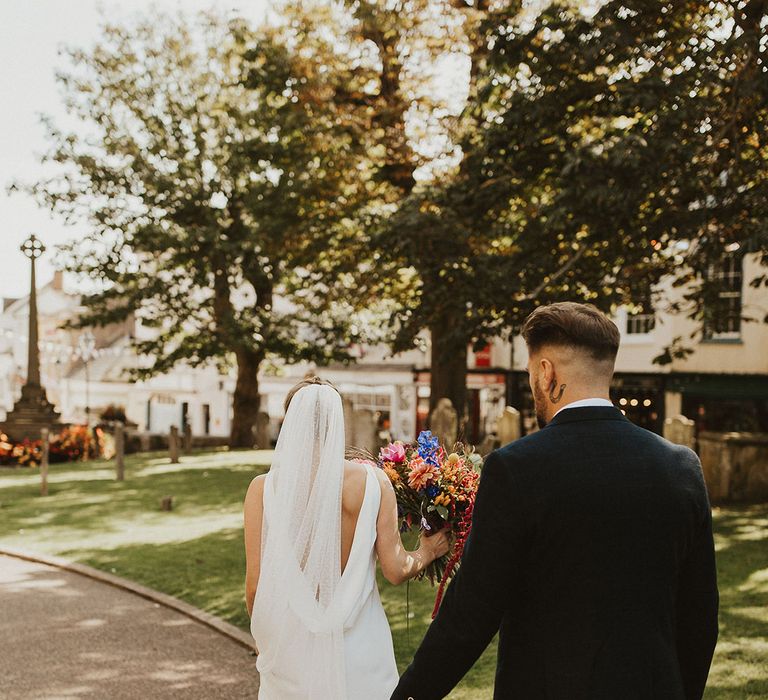 Groom in a navy suit following his bride in a cowl back slip wedding dress and cathedral length veil 