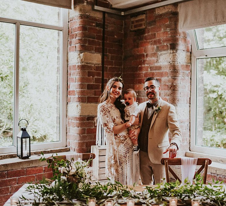 Bride & groom stand with their baby on their wedding day after ceremony