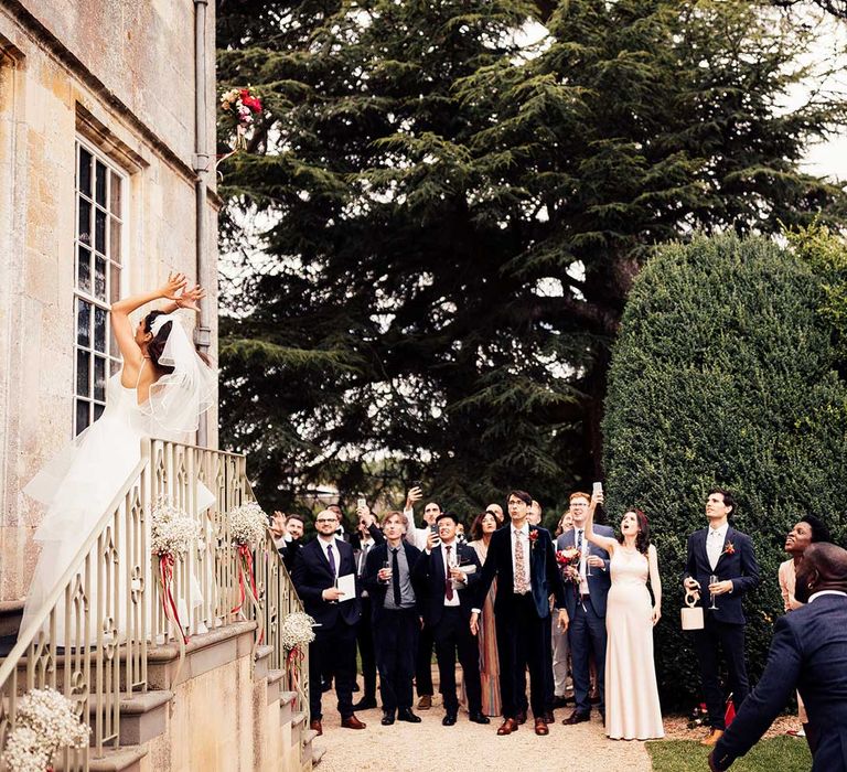 Bride in tulle skirt wedding dress and veil throws pink and red bouquet for bouquet toss into crowd of guests in the grounds of Elmore Court