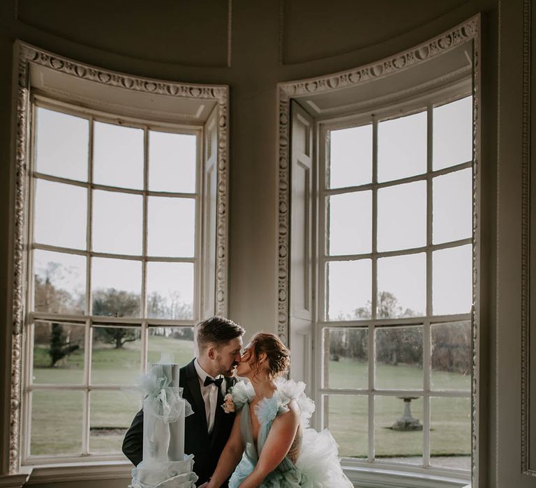 Bride in a light blue wedding dress by Millia London kissing her groom in a tuxedo with horseshoe waistcoat as their cut their monumental ruffle wedding cake 