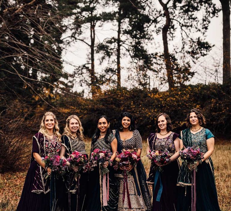 Bridesmaids in various Indian saree bridesmaid dresses all holding pink autumnal wedding bouquets with ribbon ties stand in field after wedding