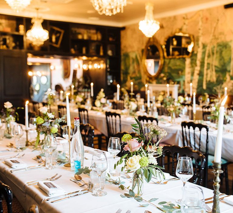 Tablescape of candles in gold antique candleholders on white tablecloth with green foliage runner