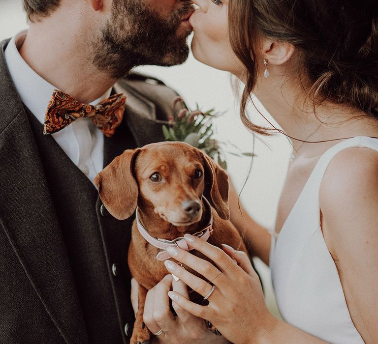 Bride and groom share a kiss with groom wearing pasiley bow tie and bride with white star nails and pearl earrings holding their dachshund pet